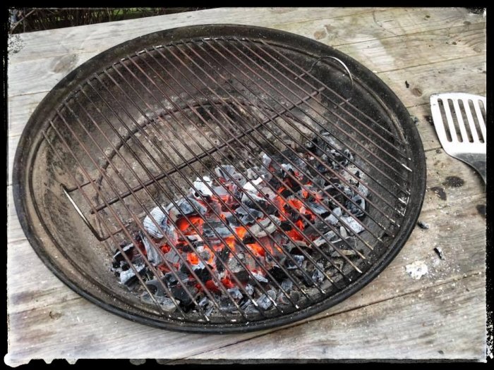 Glödande kol i en grill på en träyta med grillredskap bredvid.