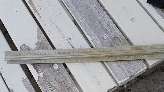 En utdragen skjutmåttstång på slitna träplankor med texten "VÄGGTJOCKLEK 1 CM".
