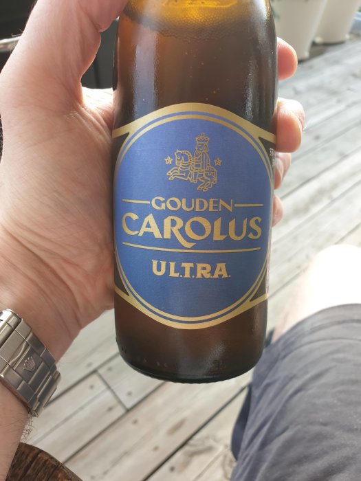 Hand som håller en flaska Gouden Carolus Ultra belgisk lättöl på en träterrass.