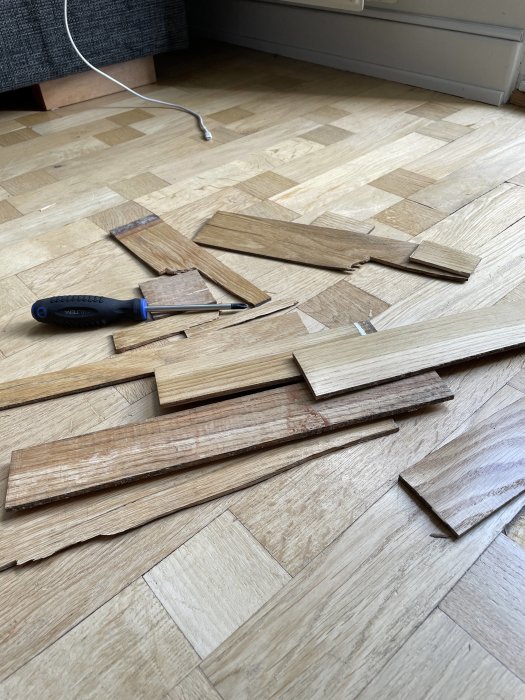 Skadat parkettgolv med uppvikta trästavar och en skruvmejsel på golvet