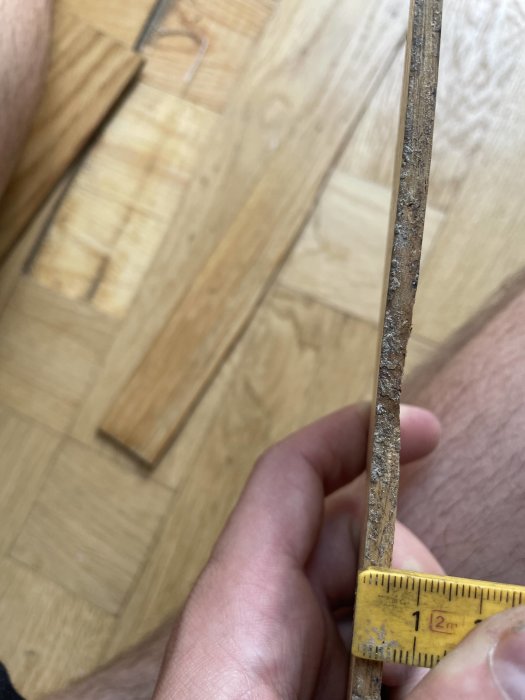 Någon mäter tjockleken på en trästav med ett måttband framför ett trägolv.