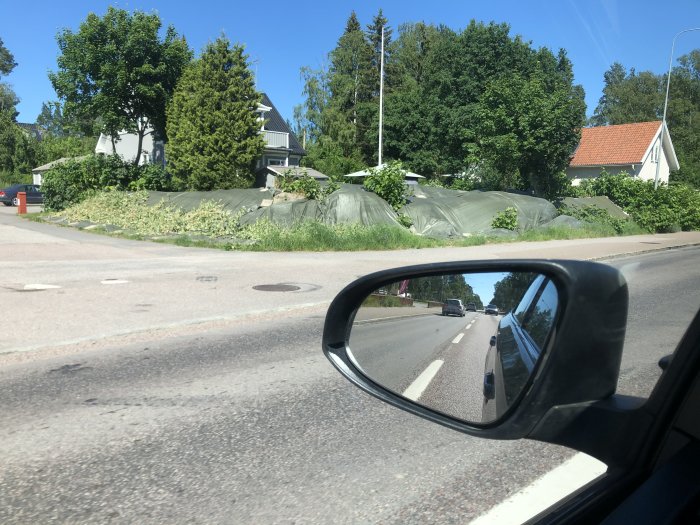 Vy genom bilfönster där vegetation täcks av presenningar vid hus, vilket antyder igångsatt bekämpning.