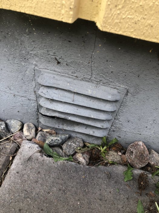 Ett grått ventilgaller i en betongvägg nära marken med småstenar framför.