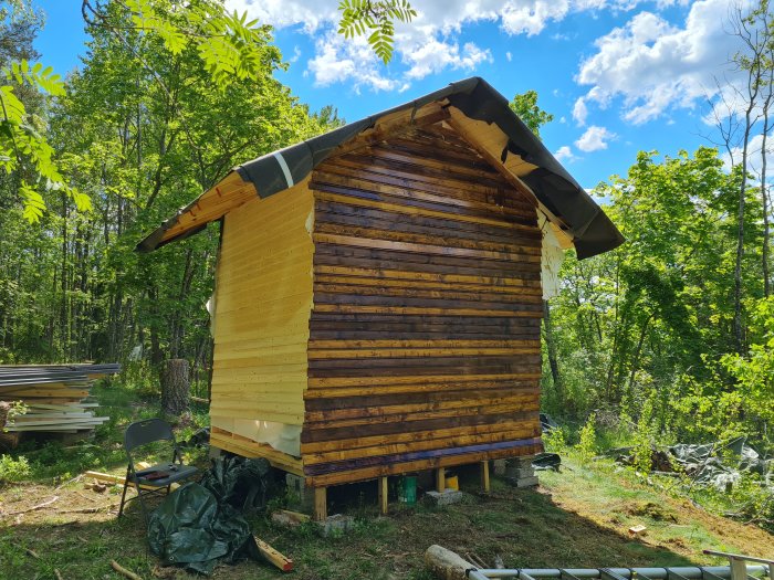 Stuga under renovering med en obehandlad främre vägg och skog i bakgrunden.