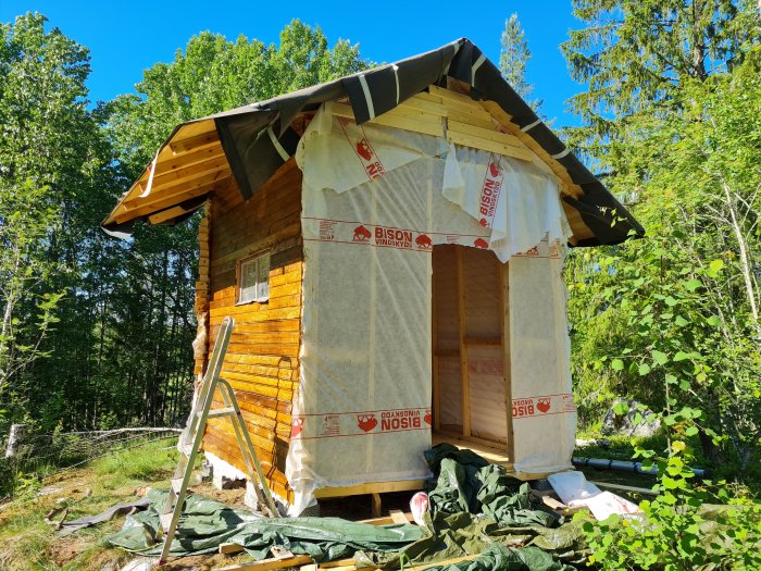 Halvfärdig stuga med tre färdiga väggar och en ofärdig främre vägg som är isolerad med vindskyddsfilt.