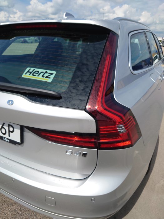 Bakre delen av en silverfärgad Volvo V60 med Hertz-dekal på bakfönstret och våt efter regn.