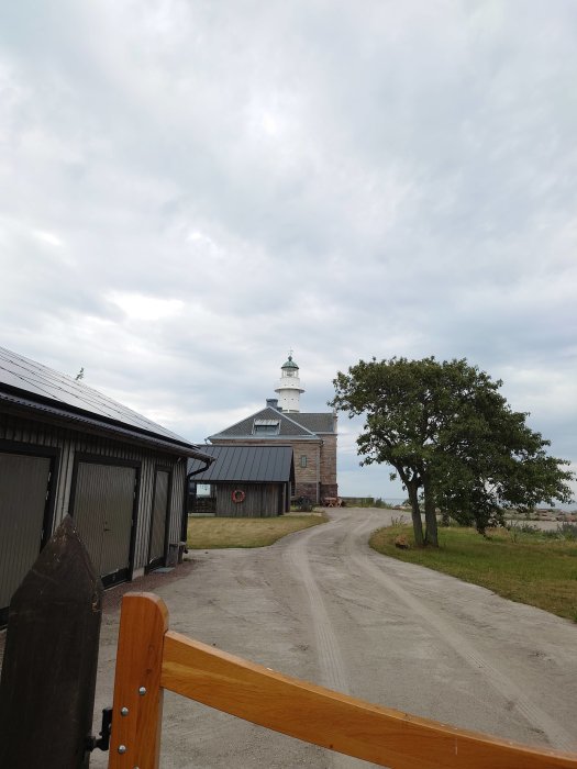 Grusväg leder till rustik byggnad med torn, omgiven av träd och staket. Molnig himmel överst.