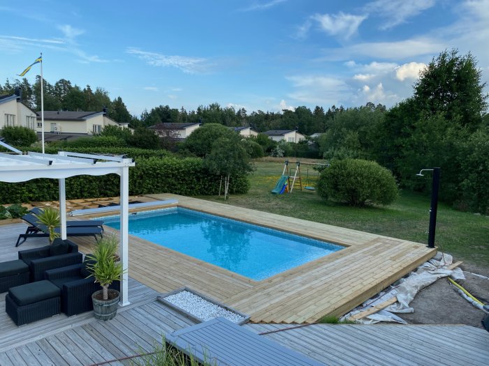 Halvfärdig trädäck runt en blå pool i en trädgård, med loungemöbler och en svensk flagga i bakgrunden.