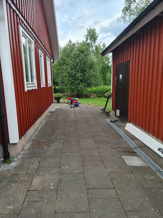 Innergård med lutar stenläggning mot ett rött hus, synlig avrinningsränna och lövträd i bakgrunden.