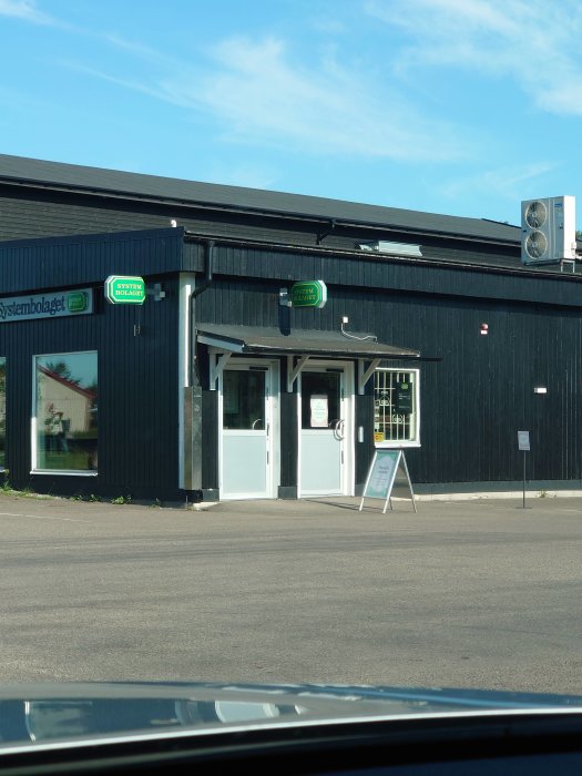 Exteriör av Systembolaget butik med svart fasad och vit entrédörr, skylt och parkering.