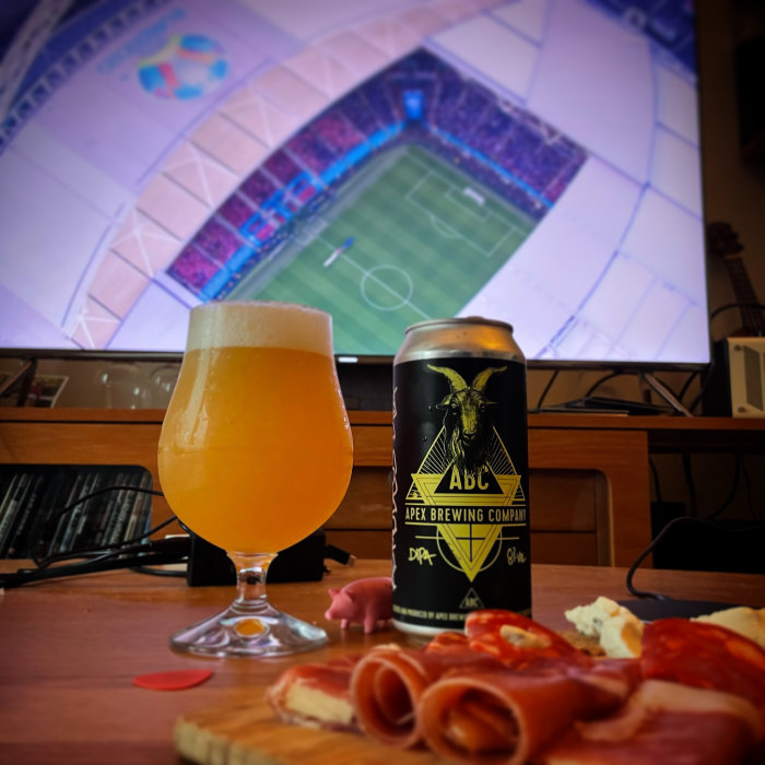 Ölglas med skum och ölburk framför TV som visar fotboll, skivad charkuteri på ett bord.