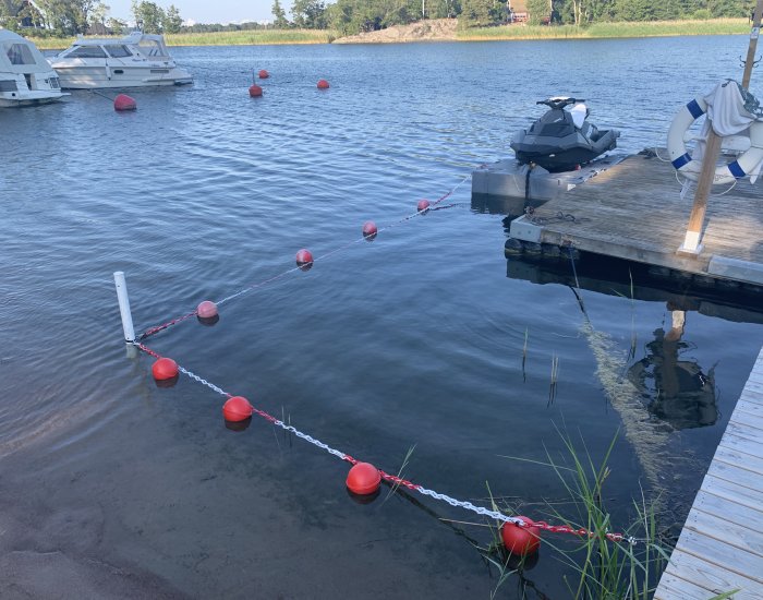 Markeringskedja med röda nätkulor vid strand för att markera djuphål nära brygga och vattenskoter.