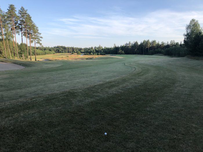 Golfbana med gräs, träd och sandbunker i förgrunden och golfboll nära betraktaren.