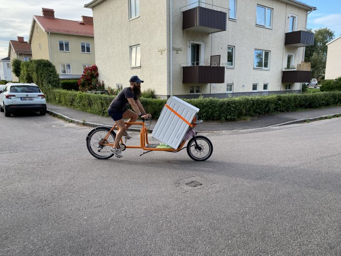 Person cyklar med en lastcykel som transporterar en stor vit låda på en stadsgata.