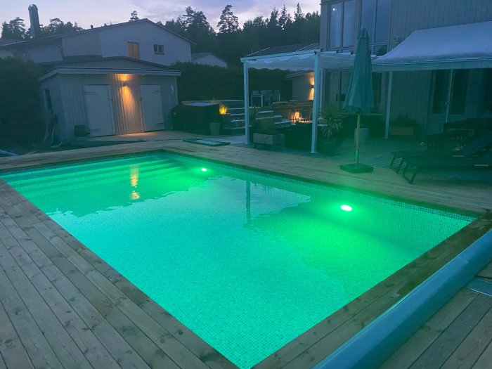 Pool med grönt undervattensljus belägen vid träaltan och hus i skymningen.