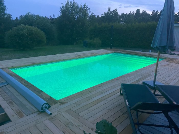 Upplyst pool med grönt ljus på träaltan under kvällshimmel, solstolar och parasoll syns i förgrunden.