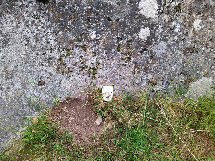 En ovanlig kubikformad sten med inskription, inbäddad i marken vid en klippvägg, omgiven av gräs och mossa.