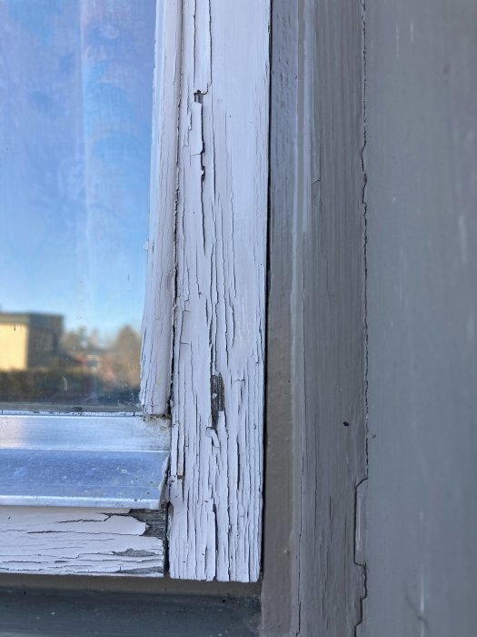 Närbild på ett gammalt fönster med flagnande, vit färg och sprickor i träet, behöver renovering.