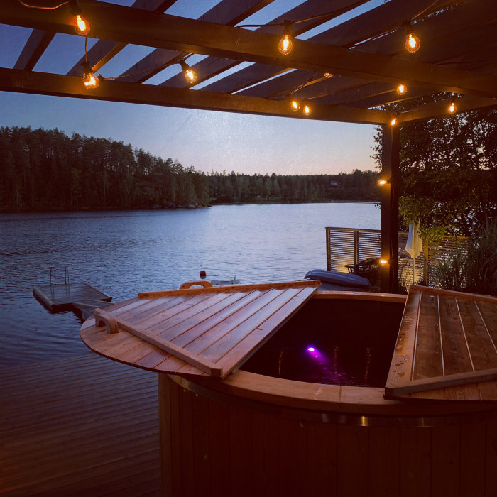 Utomhusbadtunna med öppet lock under pergola med glödlampor och utsikt över en sjö vid skymning.