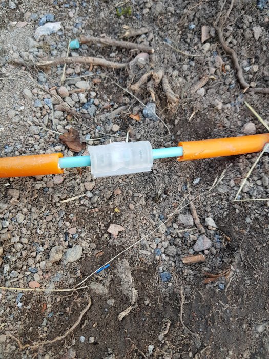 Reparerad ledning i mark med en skarvkoppling mellan två kablar omgivna av grus och småstenar.