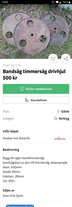 Rostiga järndrivhjul för bandsåg ligger på marken med gräs i bakgrunden.