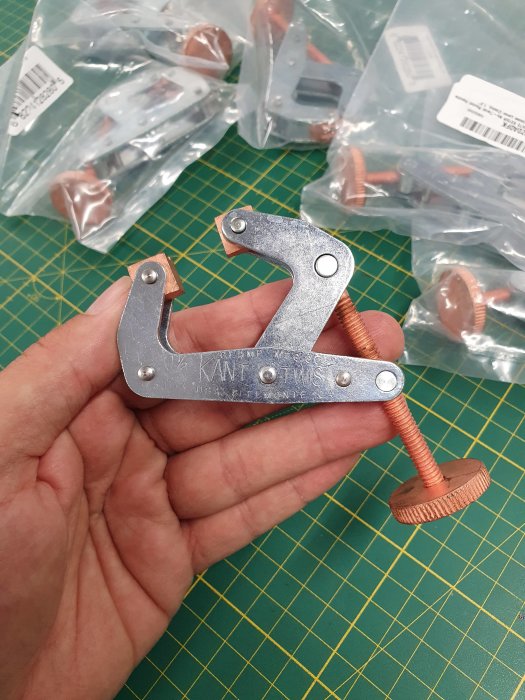 Hand håller en skruvklämma ovanför ett skrivbord med fler förpackade skruvklämmor i bakgrunden.