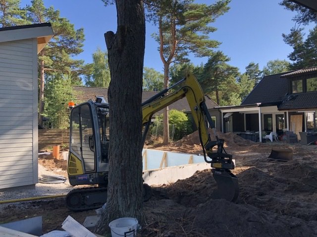 Minigrävare vid arbete i trädgård nära hus och träd, jordhögar syns.