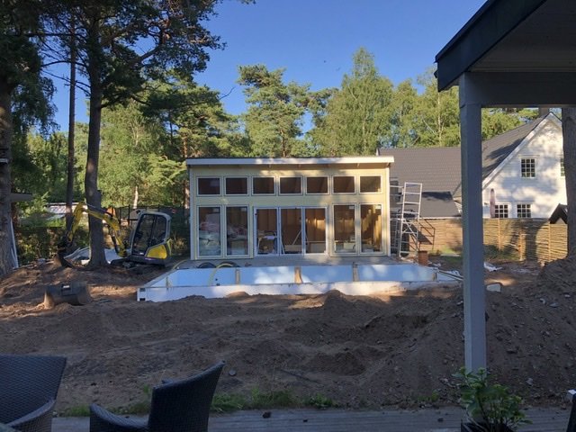 Grävmaskin och upphögar av jord vid hus med pool under konstruktion och skogsområde i bakgrunden.