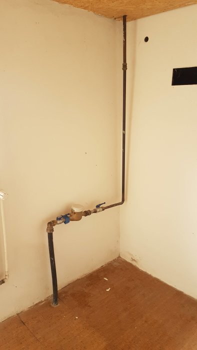 Vattenledningsrör i hörn av rum, med koppar uppåt och svart plast/gummirör nedåt, inklusive mätare och ventiler.