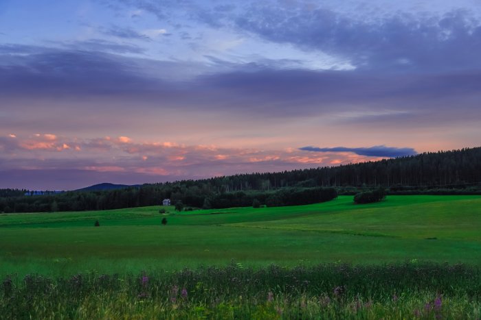 Solnedgång över lantligt landskap med gröna fält, träd och dramatisk himmel.