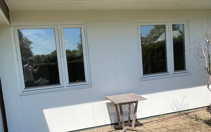 Vit yttervägg med tre fönster under takutsprång, reflektion av fotograf och trädgård i fönsterglasen.