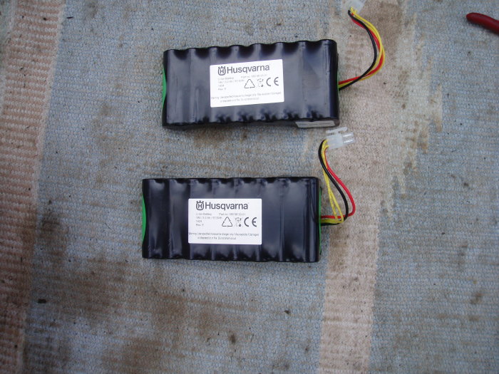 Två utbytta Husqvarna Automower batterier på ett grått underlag.