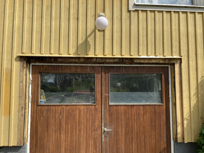 Skadade fasadbrädor med tecken på röta vid basen av ett 50-tals putshus, ovanför en brun garageport med vit smyg.