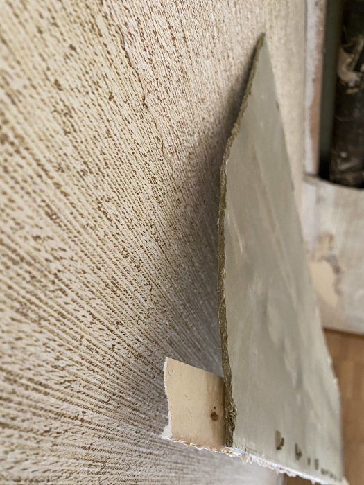 A removed panel showing a piece of board from a wall, revealing pipes behind it in a renovation project. Concerns about asbestos presence in the material.