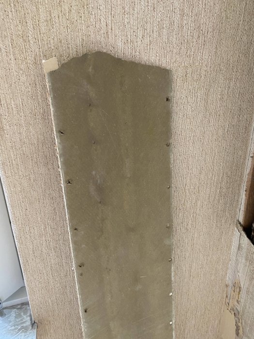 A piece of fiberboard or wall panel partially removed from an interior wall, exposing potential asbestos material, with visible screws along the edges.