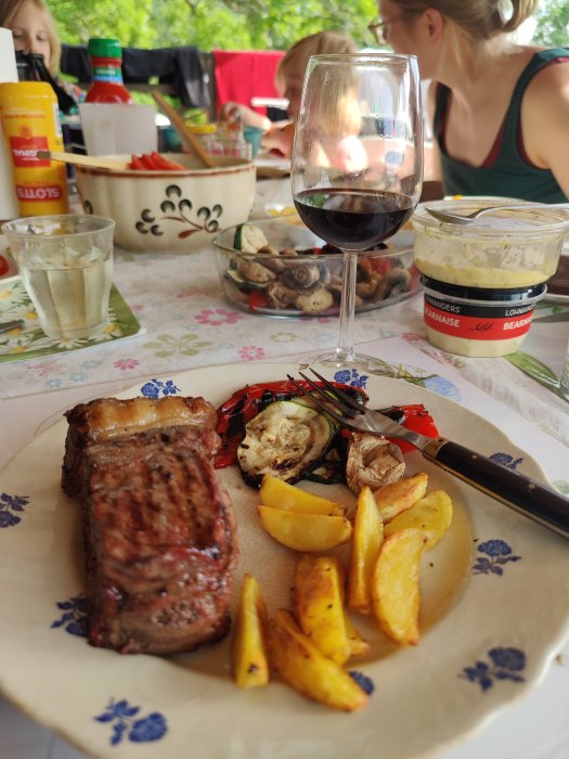 Grillad ryggbiff med kappa, klyftpotatis och grönsaker på ett dukat bord utomhus med drycker och tillbehör.