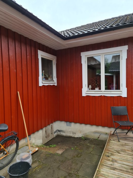 Ingång till krypgrund med synlig gjuten sula, trall och lutande plattor nära rödmålad husvägg.