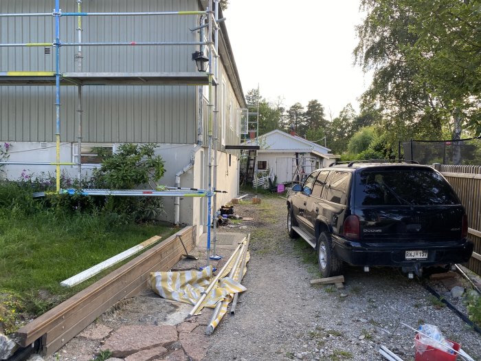 Uppfart med bil och byggmaterial framför ett hus med byggställningar och ojämn gräsyta.