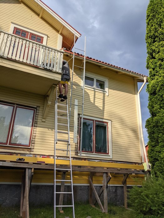 Person på en byggställning vid en gul villa med borttagen fasadsstege och delar av gammal panel.