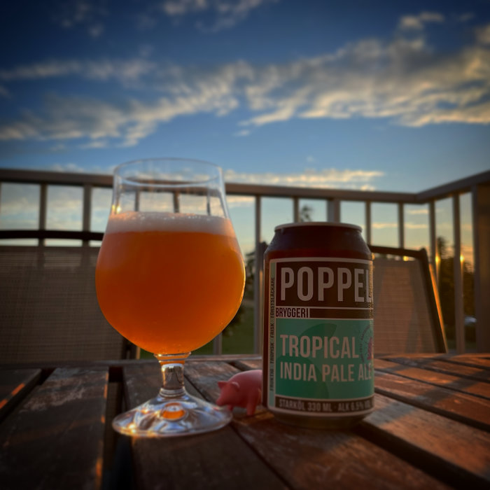 Ölglas och burk med märket "Poppel Tropical India Pale Ale" på ett träbord mot solnedgångshimmel.