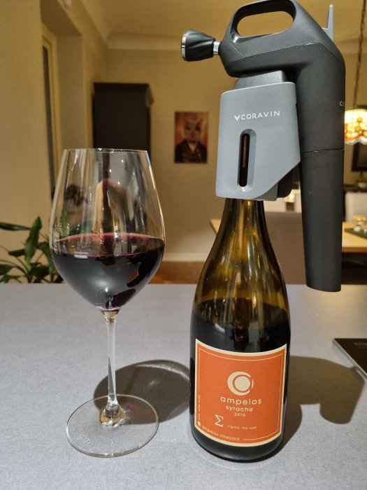 Ett glas med rödvin och en vinflaska med etiketten "Ampelos Syrache 2016" och en Coravin vinöppnare.