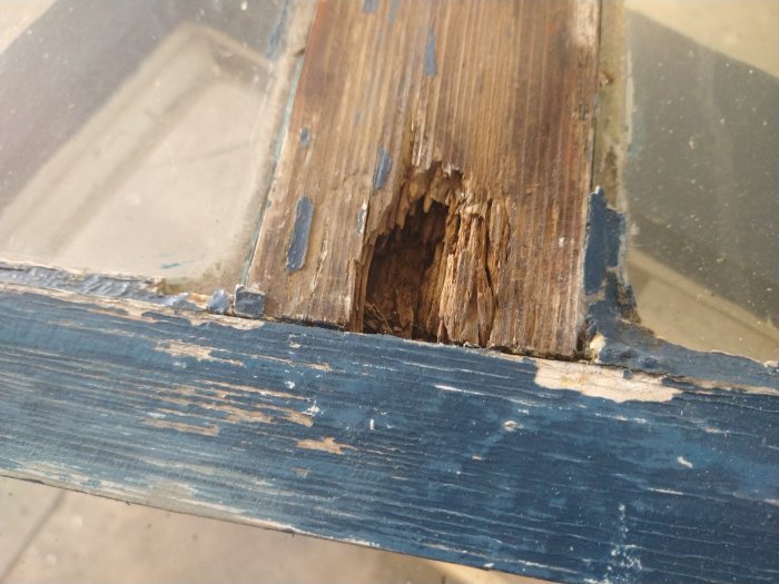 Skadat och ruttet trä på en gammal blåmålad källarfönsterbåge, behov av renovering.