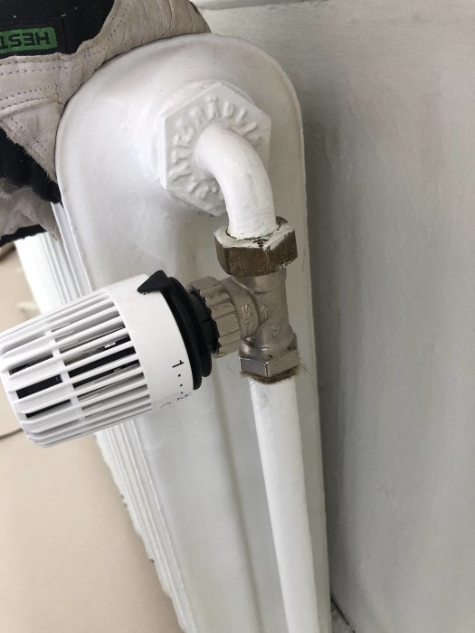 En person med en handskadad hand som håller i ett vitt radiatorrör med en termostatknopp.