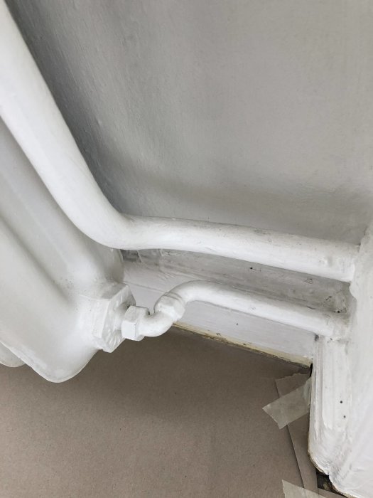 Vitmålat rörledningshörn och fot av en radiator mot en vägg, med golvskydd av tejp.