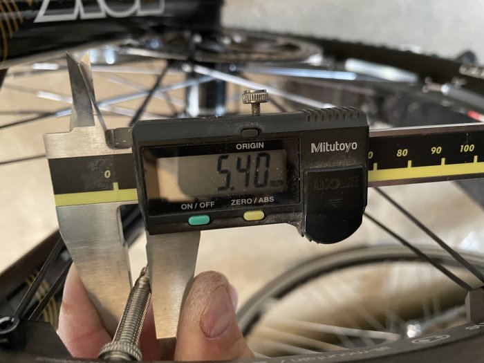 Digital skjutmått visar måttet 5,40 mm på ett cykelventilhål med cykelhjul i bakgrunden.