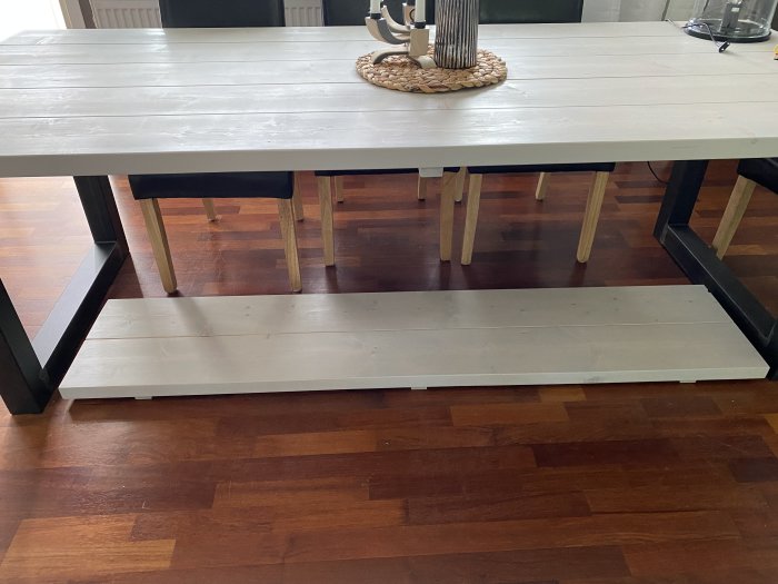 En hvitmalt benk med treplate og stålbein plassert i en stue med parkettgulv, pyntet med en rund bordbrikke med gjenstander oppå.