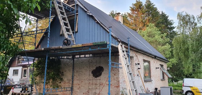 Renoverad husgavel med blå plåt och byggställning, omgiven av grönska och en stege lutad mot väggen.