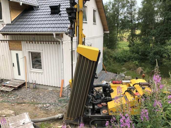 Borrutrustning för brunnar bredvid ett hus med vit fasad och skog i bakgrunden.