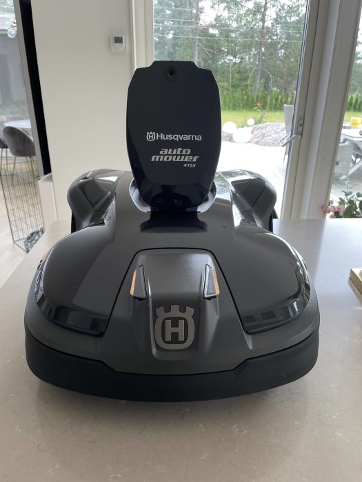 Husqvarna Automower robotgräsklippare modell 415X på ett bord inomhus med utsikt mot trädgård.