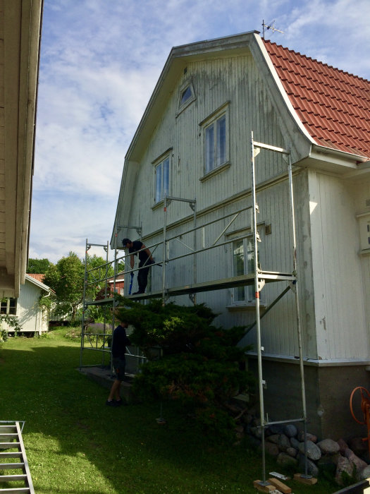 Person på byggnadsställning målar vit träfasad på bostadshus, en person assisterar nedanför.